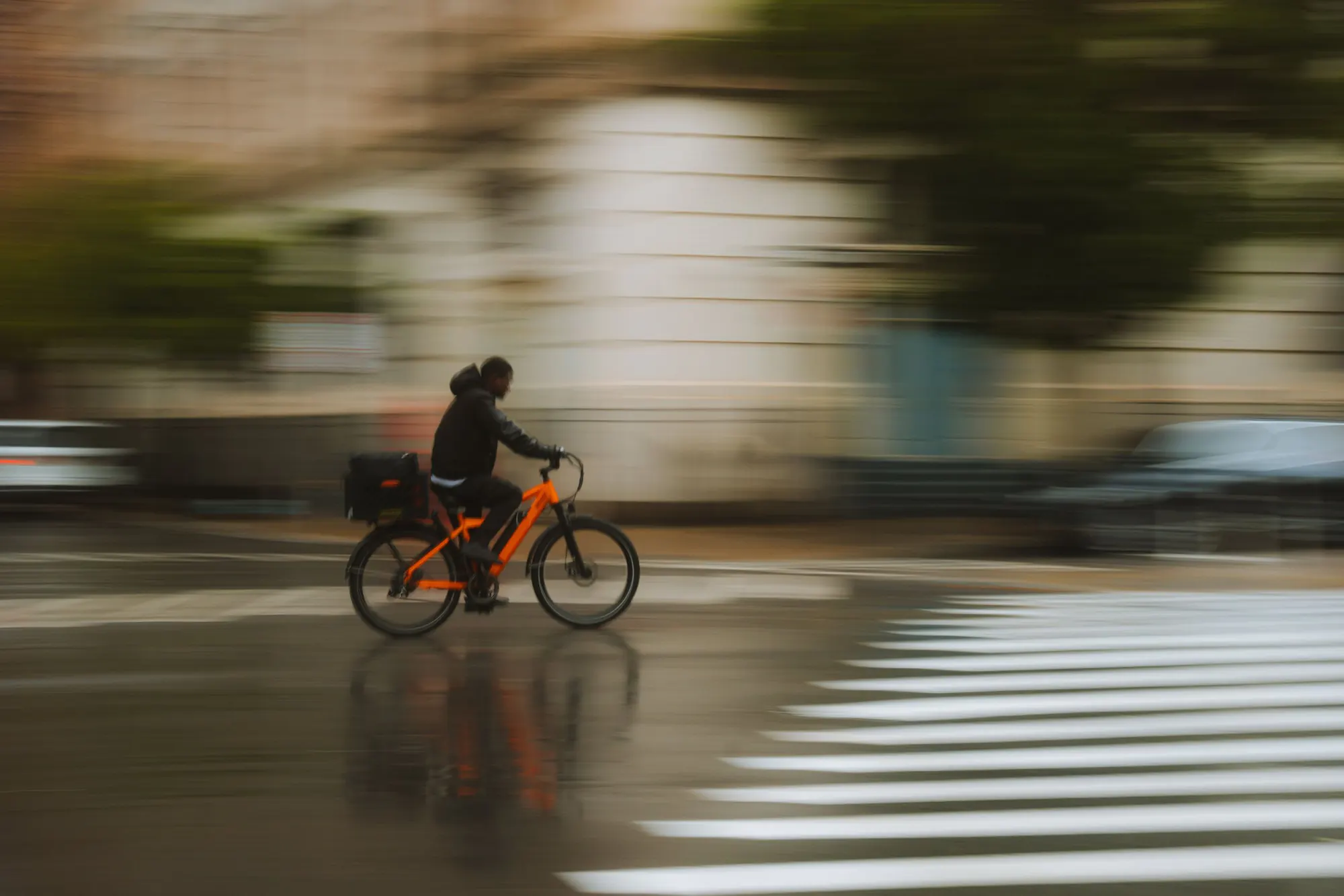 Et bud med en kasse bag på sin orange elcykel kører gennem en våd bygade i regnvejr, fotograferet med bevægelsesslør