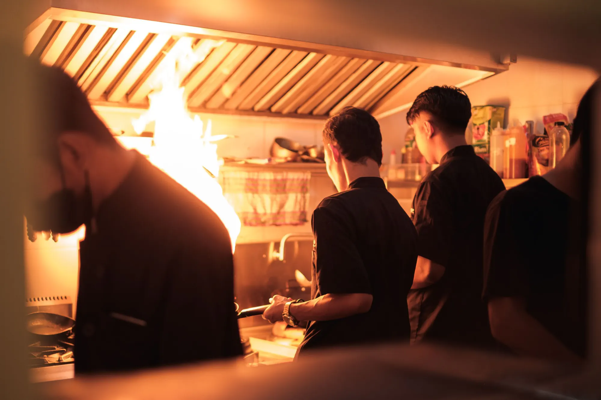 Kokke i sort arbejder ved komfurerne i et travlt restaurantkøkken, hvor en stikflamme lyser op under emhætten