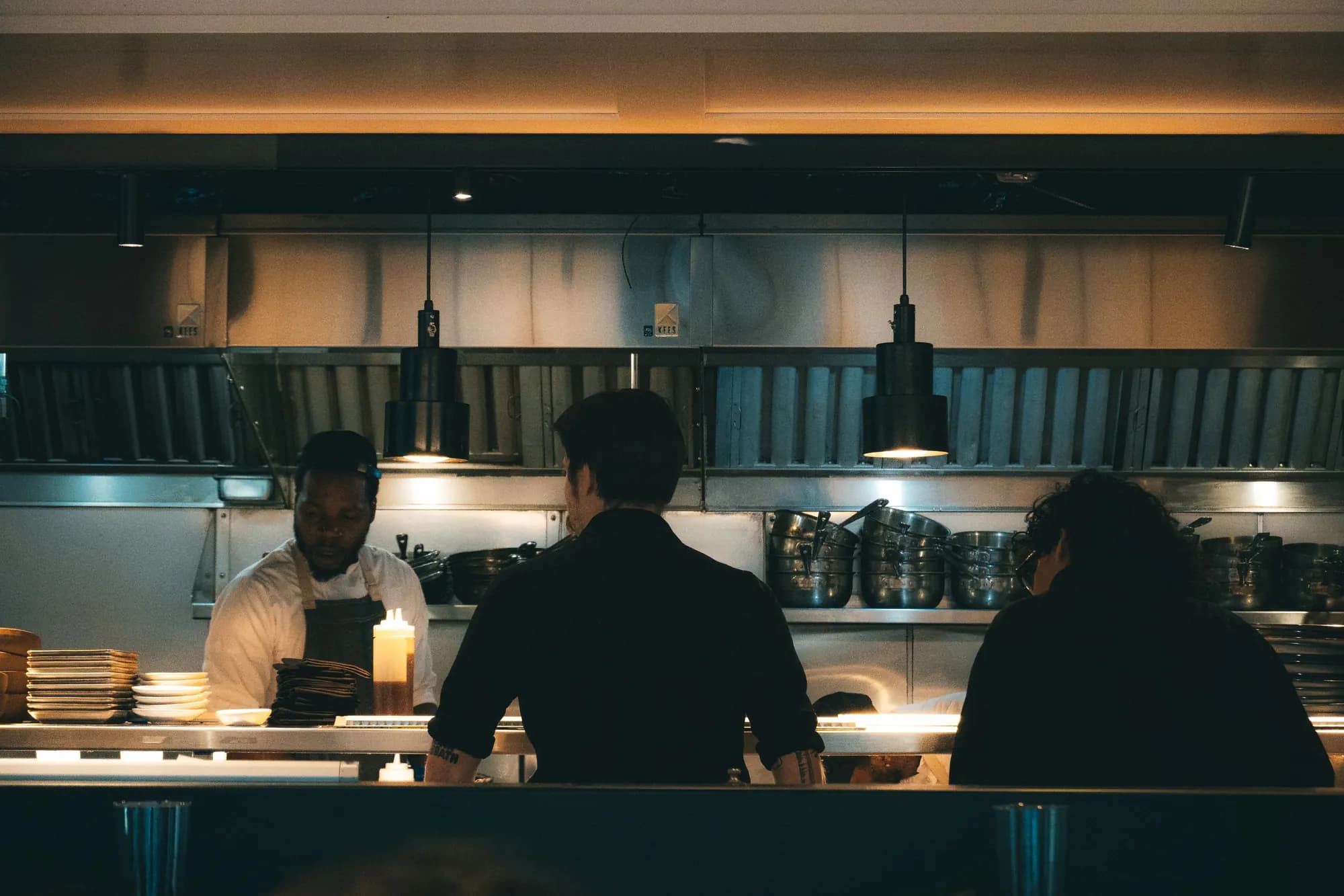 Kig gennem passet ind i et restaurantkøkken om aftenen, hvor kokkene arbejder under hængelamper