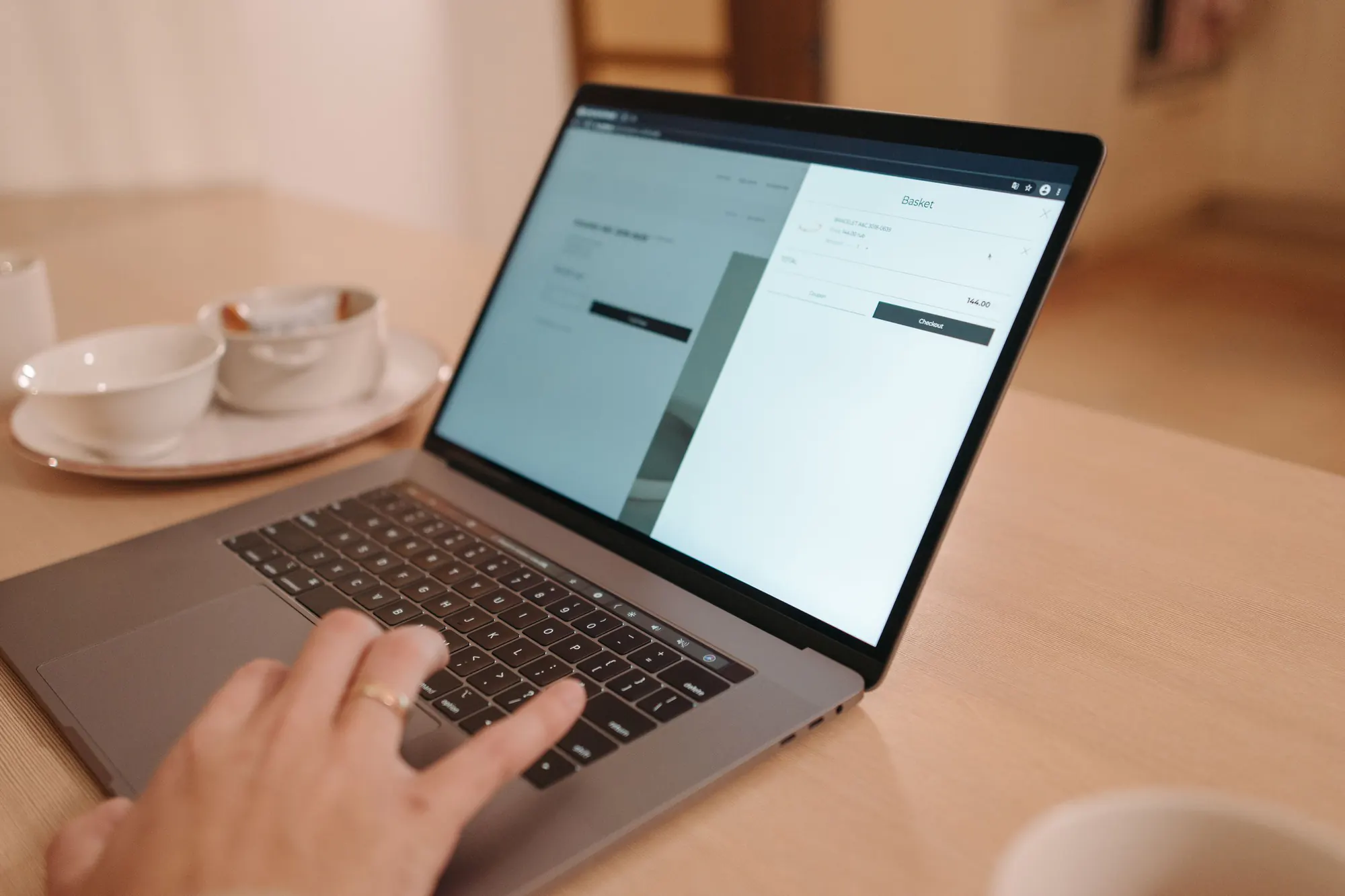 En hånd på tastaturet til en bærbar computer med en webshops indkøbskurv på skærmen ved et lyst træbord med kaffekopper