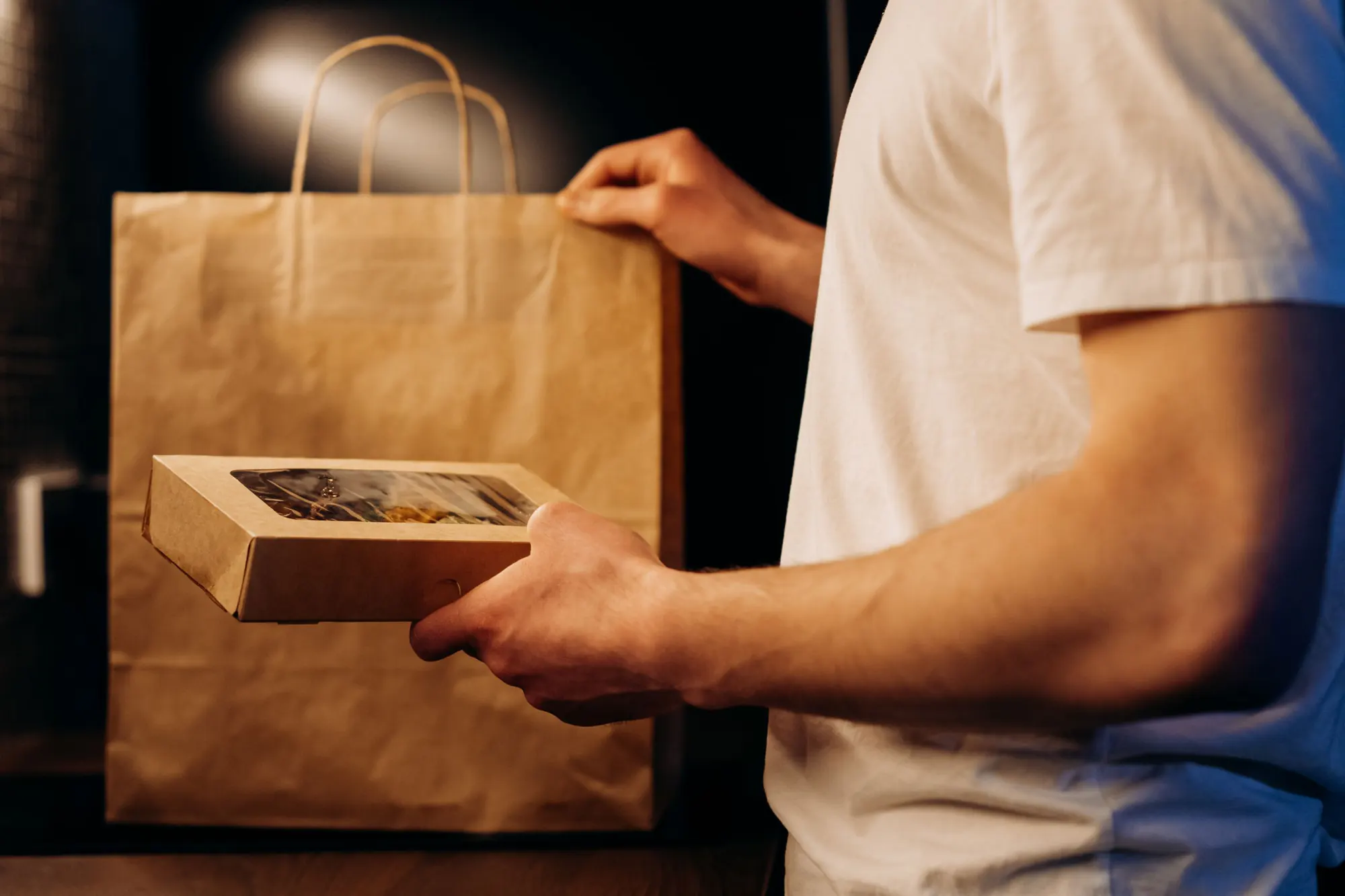 En hånd holder en brun take away-boks med rude op foran en papirpose, klar til afhentning