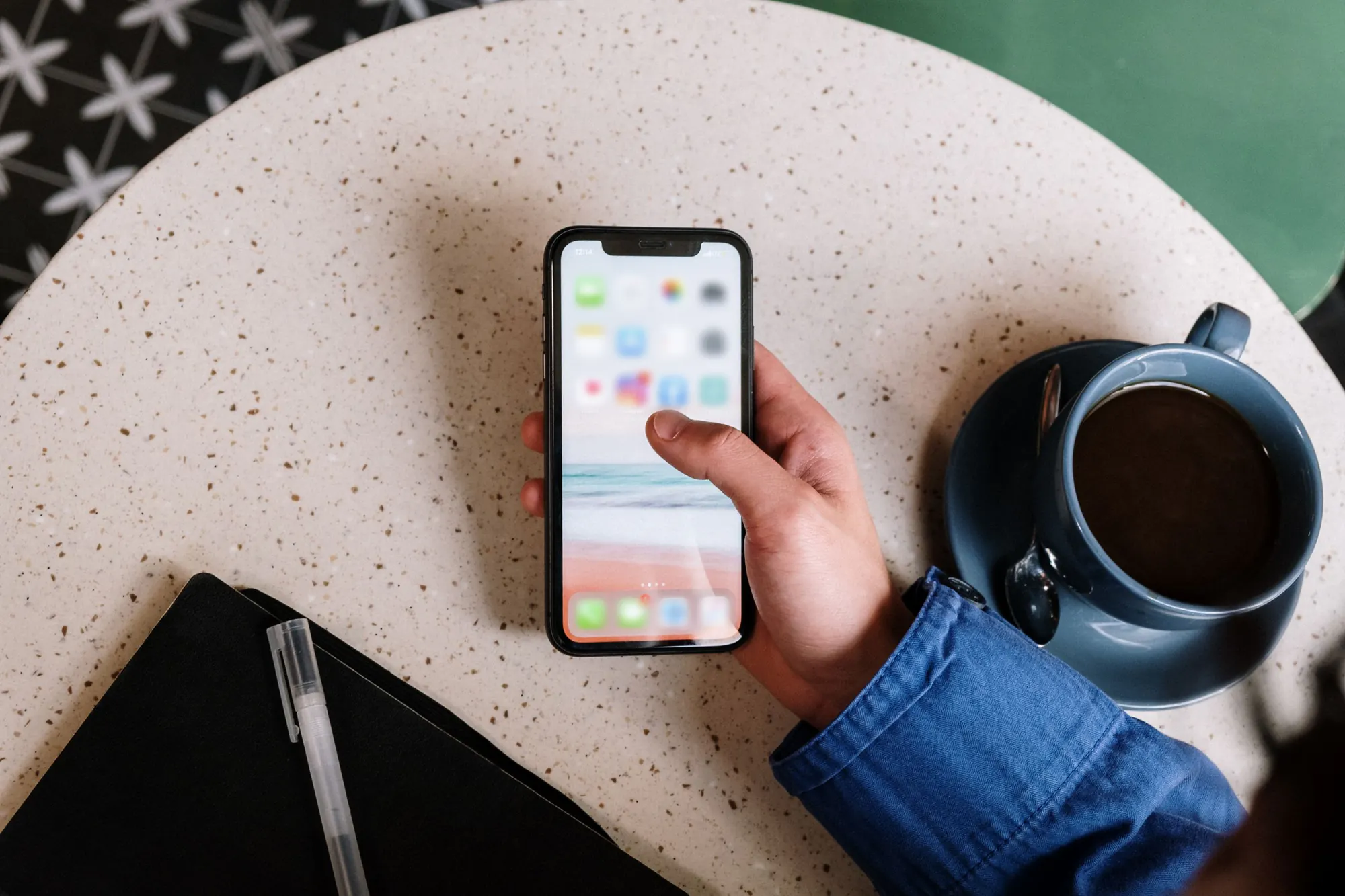 En hånd holder en telefon med app-ikoner på hjemmeskærmen over et cafébord i terrazzo ved siden af en kop sort kaffe