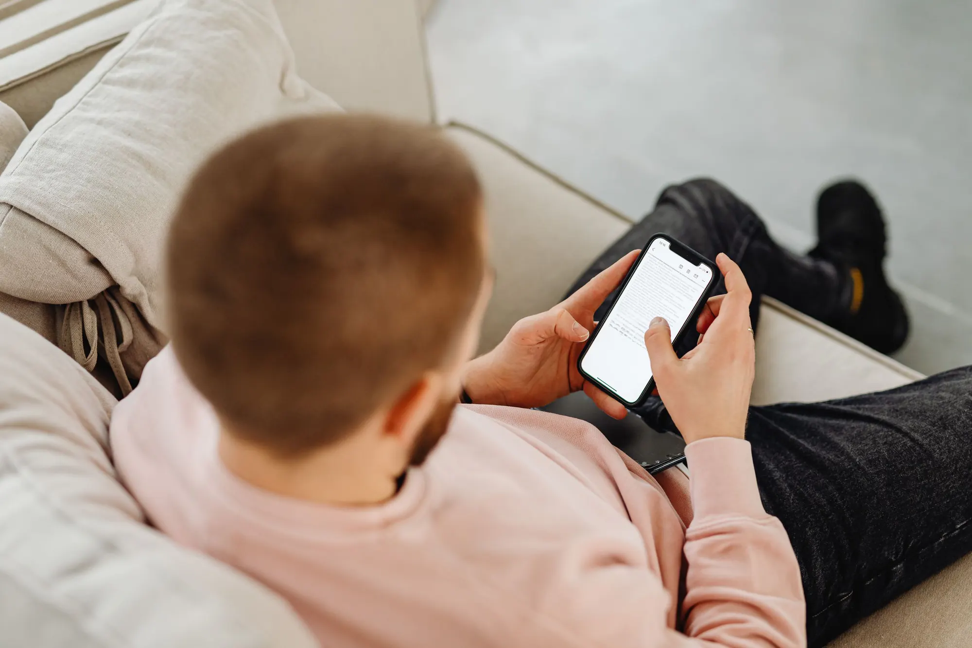 En mand i lyserød sweater ligger i en lys sofa og bladrer på sin telefon, set bagfra
