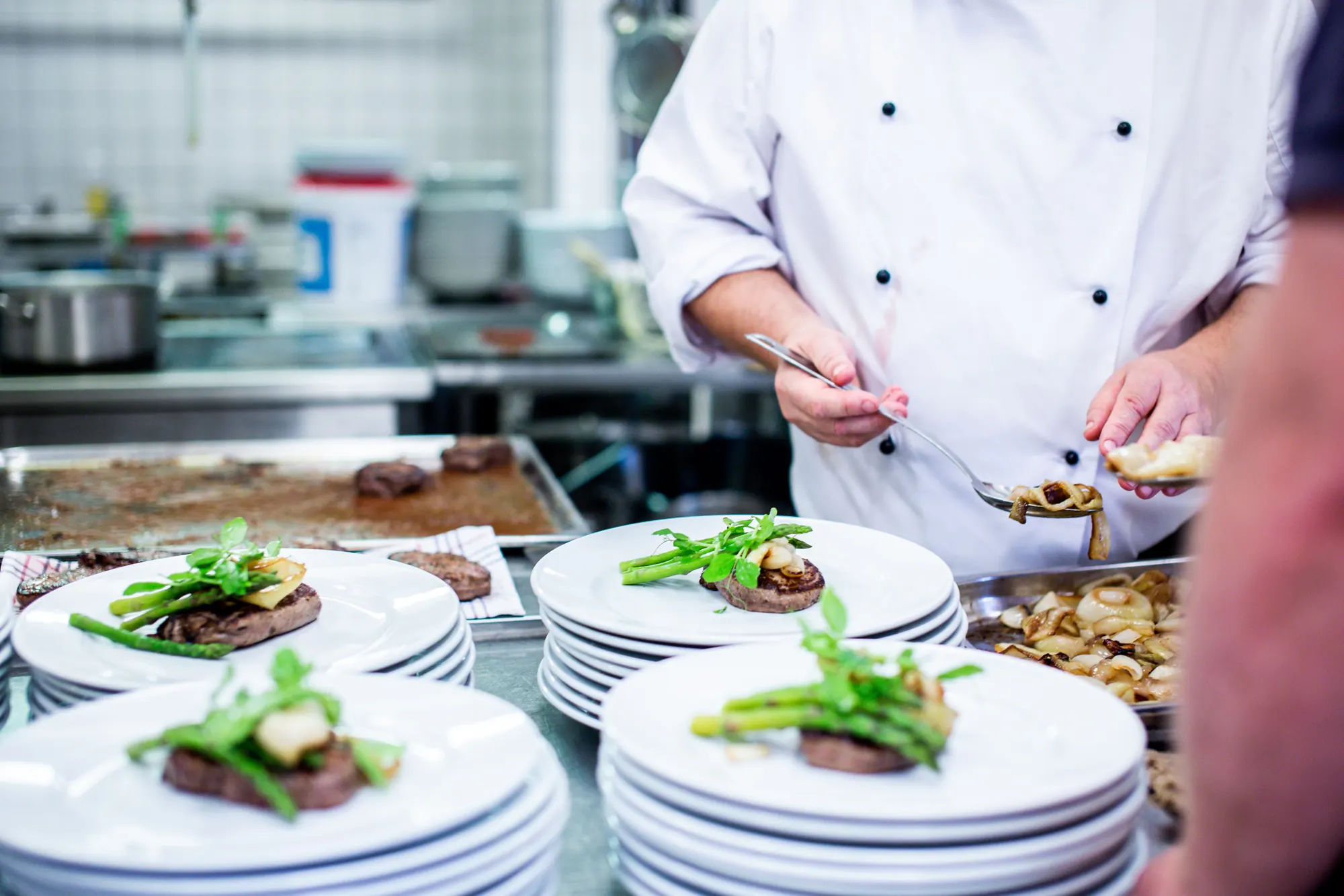 En kok i hvid kokkejakke anretter bøf med grønne asparges på stakke af hvide tallerkener i et travlt restaurantkøkken